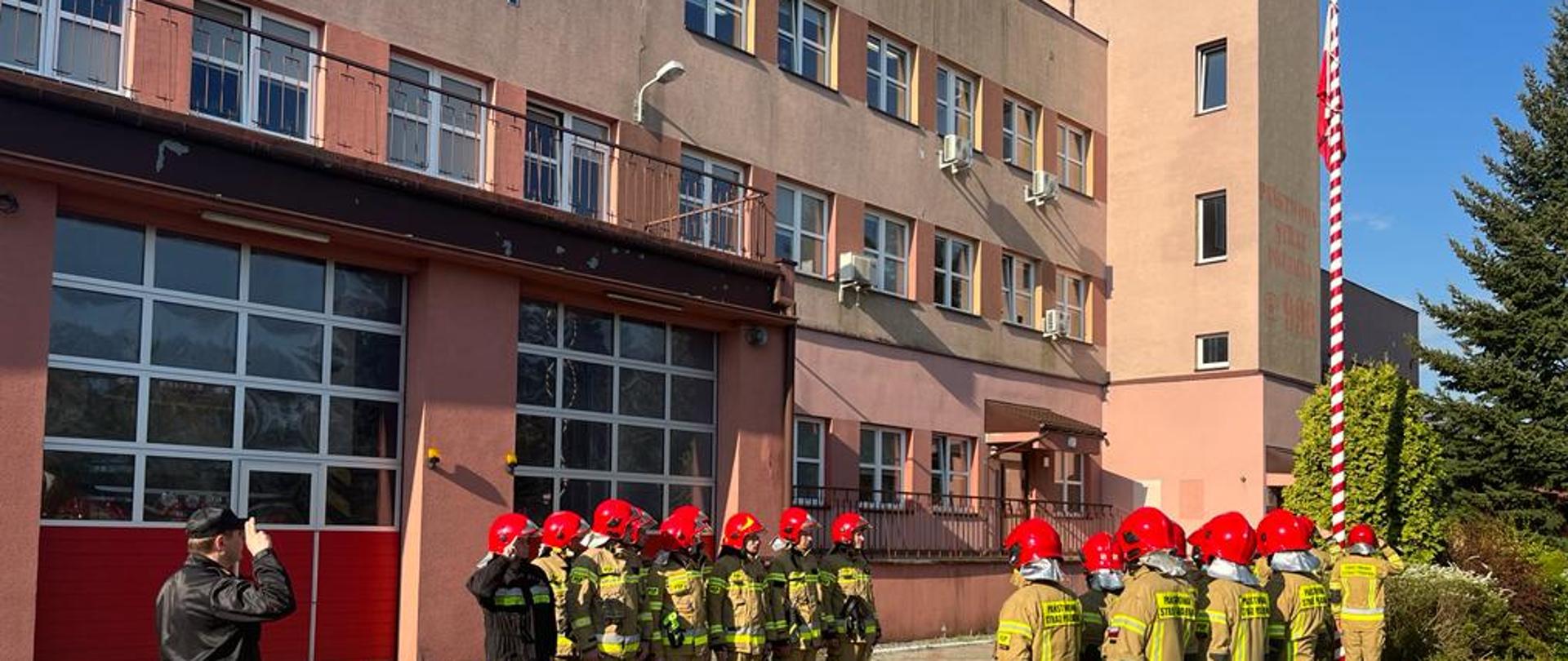 na zdjęciu strażacy w szyku w ubraniu bojowym z czerwonymi hełmami w trakcie obchodów Dnia flagi ,dalej trzech strażaków podnosi flagę na maszt w tle budynek straży