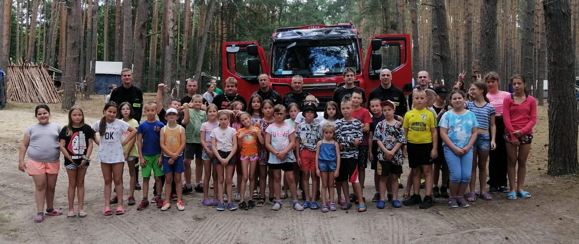 Zdjęcie zrobione w lesie, na terenie obozu harcerskiego. W tle widać samochód strażacki, przed którym ustawiona jest grupa harcerzy wraz ze strażakami i opiekunami.