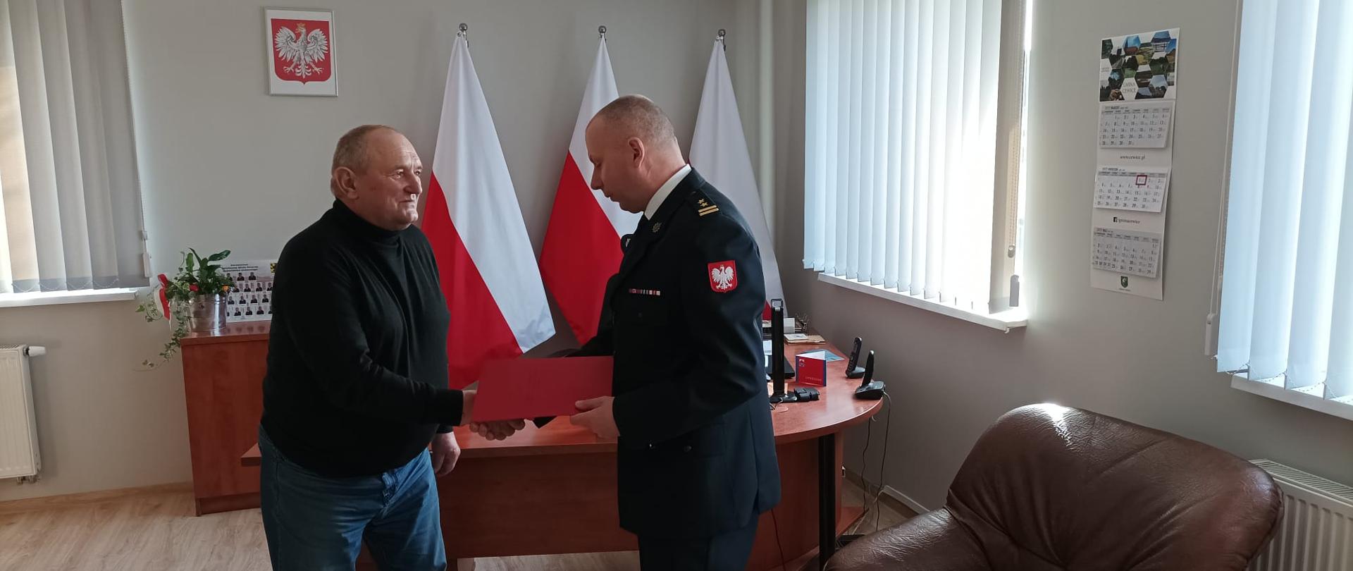 Zdjęcie przedstawia Komendanta Powiatowego Państwowej Straży Pożarnej w Lęborku wręczającego świadczenie ratownicze strażakowi Ochotniczej Straży Pożarnej. Świadczenie wręczane jest w gabinecie komendanta. W tle widoczne godło państwowe oraz trzy flagi państwowe.
