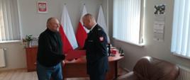 Zdjęcie przedstawia Komendanta Powiatowego Państwowej Straży Pożarnej w Lęborku wręczającego świadczenie ratownicze strażakowi Ochotniczej Straży Pożarnej. Świadczenie wręczane jest w gabinecie komendanta. W tle widoczne godło państwowe oraz trzy flagi państwowe.