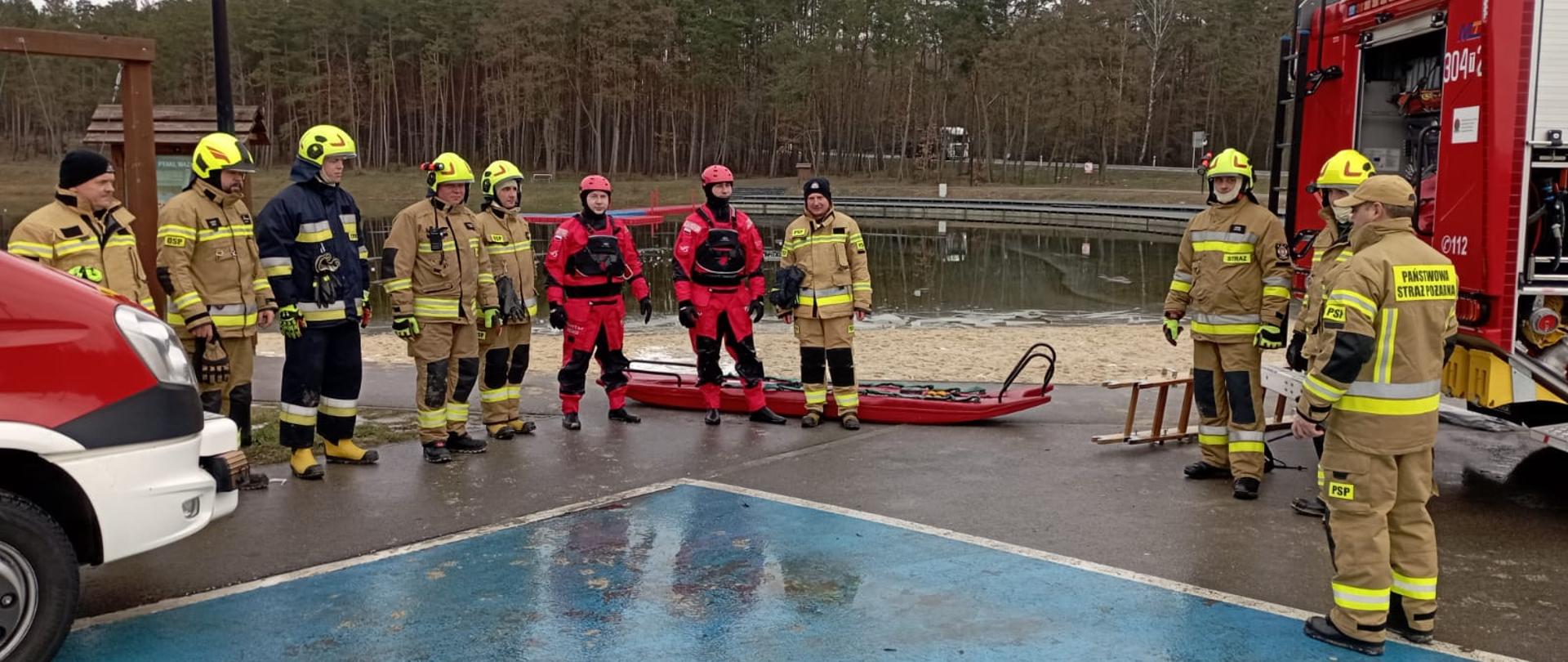 Zdjęcie przedstawia uczestników ćwiczeń stojących na parkingu przy zalewie. Za stojącymi widać zalew i las. Przy ratownikach przygotowane są sanie lodowe oraz przęsła drabiny.