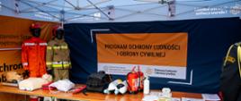 Fotografia przedstawia stoisko informacyjno-edukacyjne przygotowane pod białym namiotem ekspozycyjnym, poświęcone tematyce ochrony ludności i obrony cywilnej.