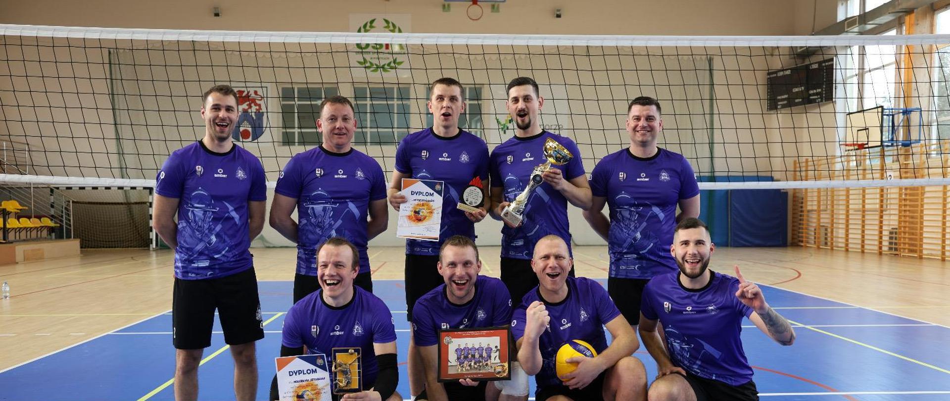 Grupa dziewięciu mężczyzn w niebieskich koszulkach sportowych pozuje do zdjęcia na hali sportowej przy siatce do siatkówki. Część z nich trzyma puchary, dyplomy i statuetki, a jeden zawodnik trzyma piłkę do siatkówki. Drużyna wygląda na zwycięską i uśmiecha się do zdjęcia.