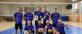 Grupa dziewięciu mężczyzn w niebieskich koszulkach sportowych pozuje do zdjęcia na hali sportowej przy siatce do siatkówki. Część z nich trzyma puchary, dyplomy i statuetki, a jeden zawodnik trzyma piłkę do siatkówki. Drużyna wygląda na zwycięską i uśmiecha się do zdjęcia.