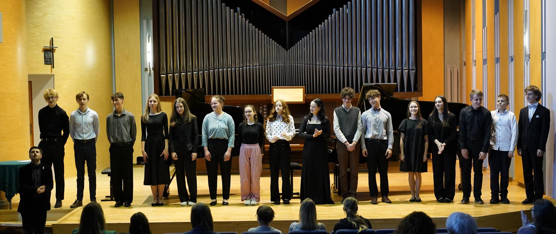 Zdjęcie grupowe. Wszyscy wykonawcy koncertu stoją w jednym rzędzie na scenie sali koncertowej, za nimi fortepian oraz organy, na pierwszym planie widownia i kilka pustych foteli