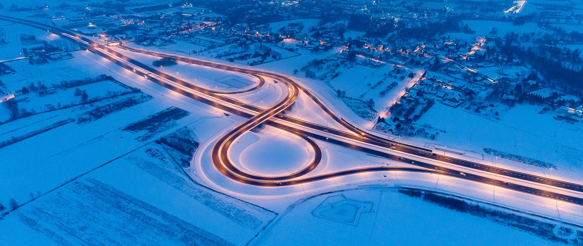 Zimowe zdjęcie z drona węzła na autostradzie A4. Jest to węzeł Łańcut wieczorną porą, widać oświetloną drogę. Droga odśnieżona. 