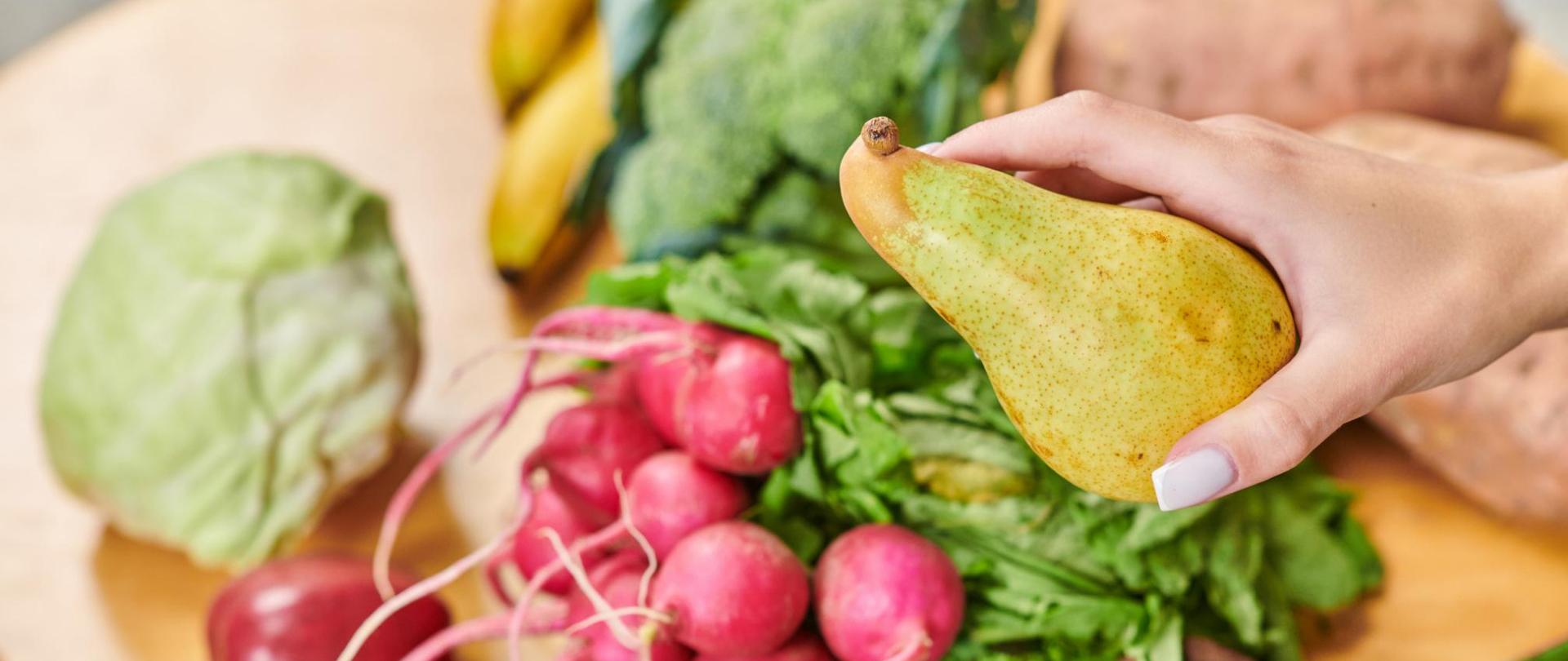 Na zdjęciu na pierwszym planie widać dłoń kobiety, w której trzyma gruszkę. W tle na blacie leżą warzywa i owoce.