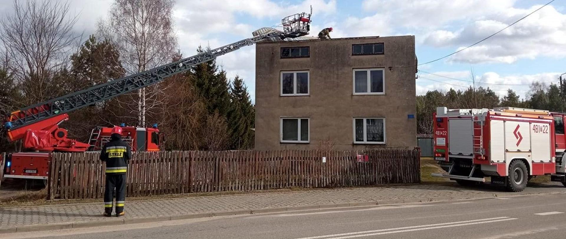 Zdjęcie obrazuje skarżyskich strażaków podczas usuwania skutków silnych wiatrów. Z lewej strony zdjęcia widzimy rozłożoną drabinę, po której strażacy dostali się na budynek mieszkalny. Znajduje się on w centralnej części zdjęcia. Na budynku strażak usuwa naderwane poszycie dachową (blachę). Z prawej strony zdjęcia strażacki wóz zabezpiecza wjazd na posesję. 