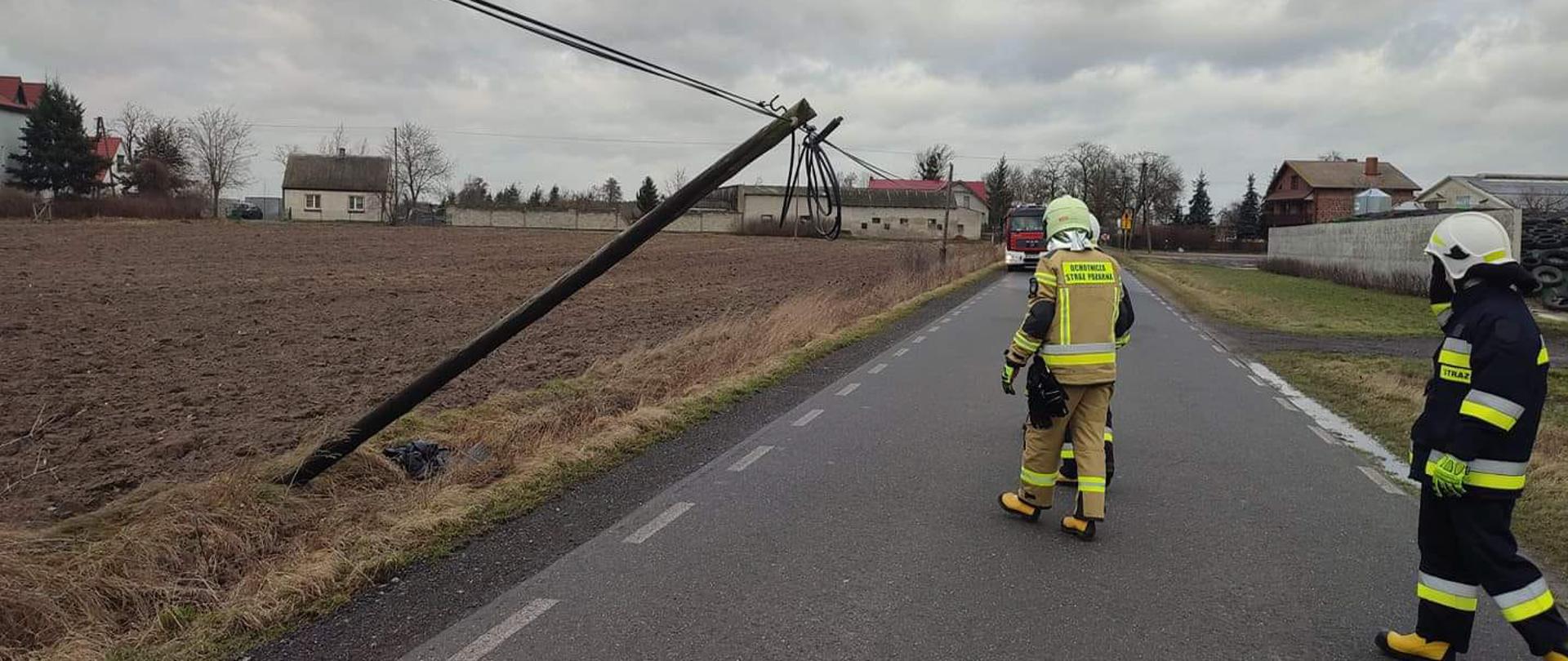 Zdjęcie przedstawia przechylony słup telefoniczny nad drogą oraz trzech strażaków