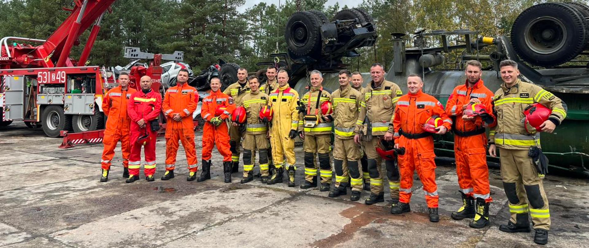 Grupa strażaków stoi obok dźwigu, wszyscy pozują do zdjęcia. Ubrani są w piaskowe i pomarańczowe mundury. W tle widać zielony las i błękitne niebo.