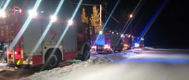 cztery pojazdy pożarnicze ustawione na drodze biegnącej z lewego dolnego rogu zdjęcia do środkowej cześć prawej strony. Pora nocna, pojazdy oświetlone.