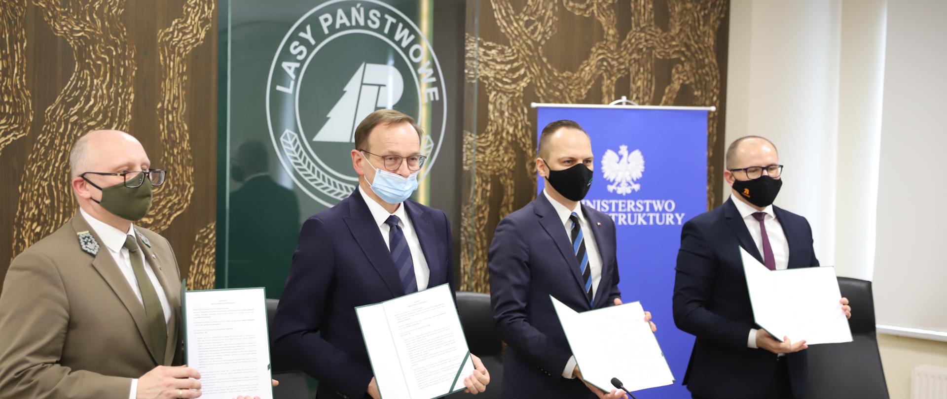 Na zdjęciu widać 4 mężczyzn którzy stoją i trzymają w rękach dokumenty. Mają ciemne garnitury i jasne koszule. W ich tle jest brązowa ściana. 