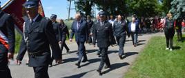 Zawody sportowo-pożarnicze. Drogą wiodącą na stadion w Sarnowie maszeruje korowód. Za pocztami sztandarowymi idą goście. Wśród nich jest m.in. komendant powiatowy PSP wraz z zastępcą, za nimi goście oraz zawodnicy.Dookoła zieleń. 