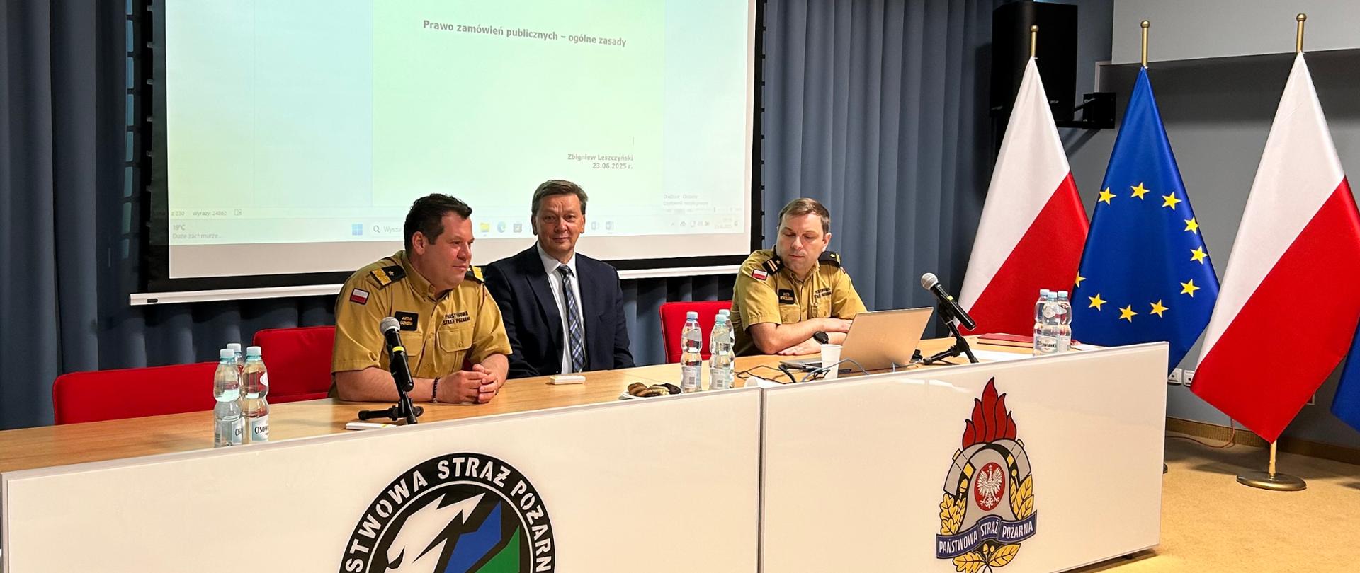 Dwóch strażaków i jeden mężczyzna w garniturze siedzi za stołem. Za plecami siedzących wisi duży biały ekran. obok, po prawej stronie stoją flagi Polski i Unii Europejskiej. 
