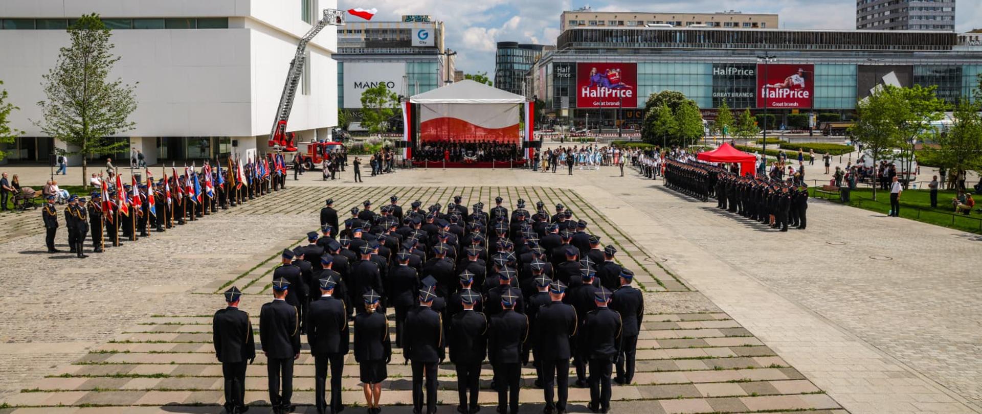 Widok z góry na plac przed Pałacem Kultury i Nauki podczas Mazowieckich Obchodów Dnia Strażaka 2026 . Na placu ustawione są pododdziały PSP, poczty sztandarowe oraz scena główna z uczestnikami uroczystości.