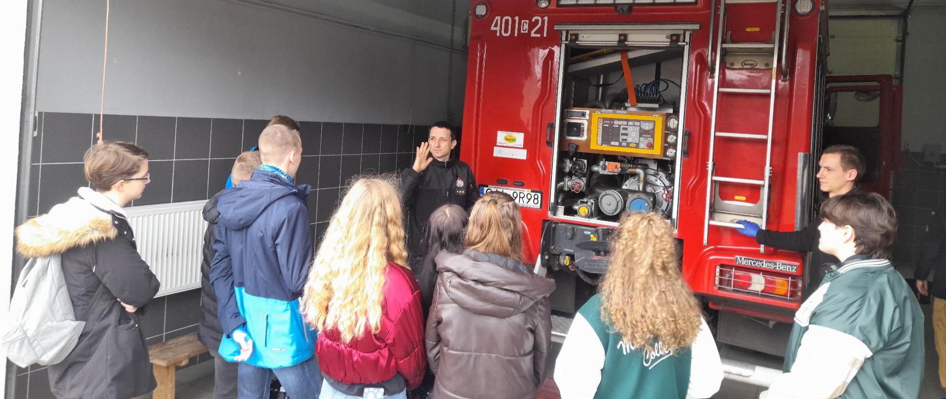 młodzież w różnokolorowych kurtkach oraz strażaka w czarnym ubraniu koszarowym który opowiada co znajduje się w skrytkach samochodu stojącej młodzieży przy samochodzie pożarniczym który jest koloru czerwonego 