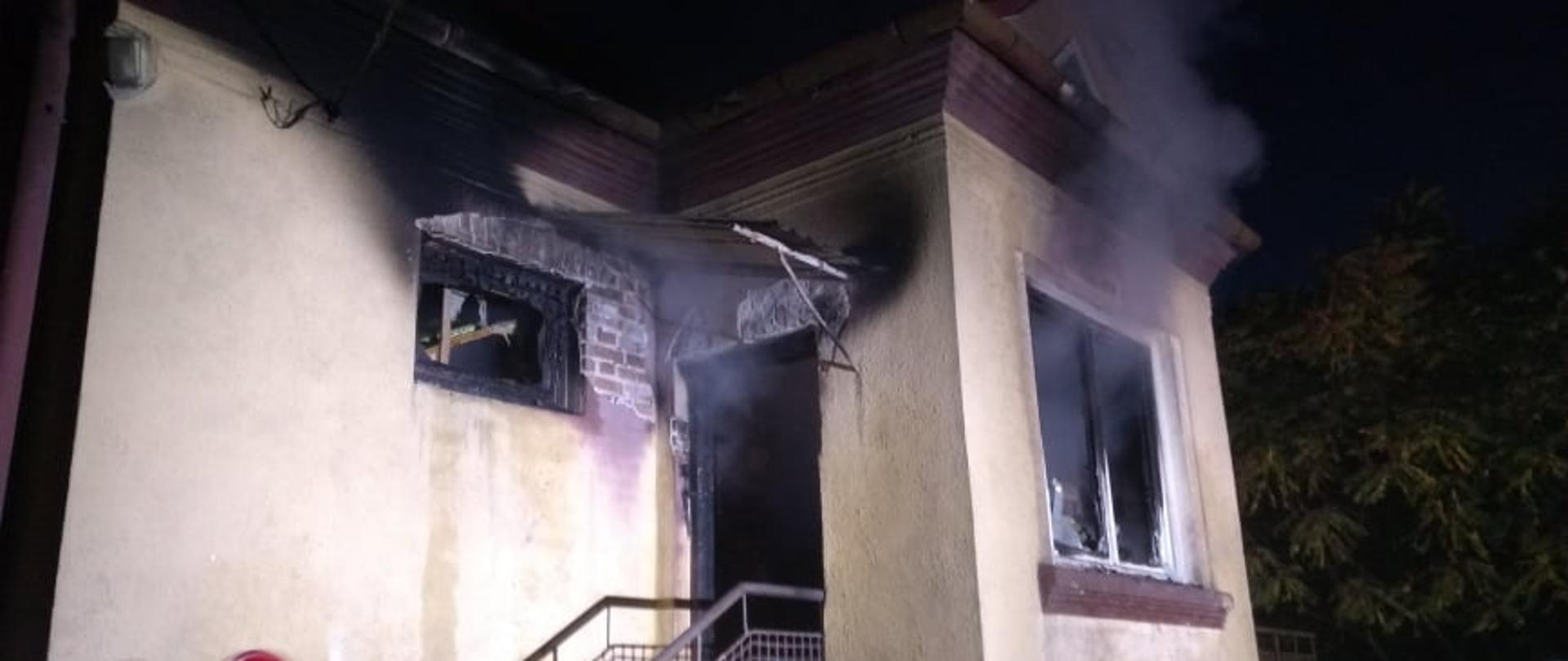 Na zdjęciu widoczna część budynku mieszkalnego. Z drzwi i okien wydobywa się dym. Na pierwszym planie widoczni częściowo strażacy prowadzący działania.