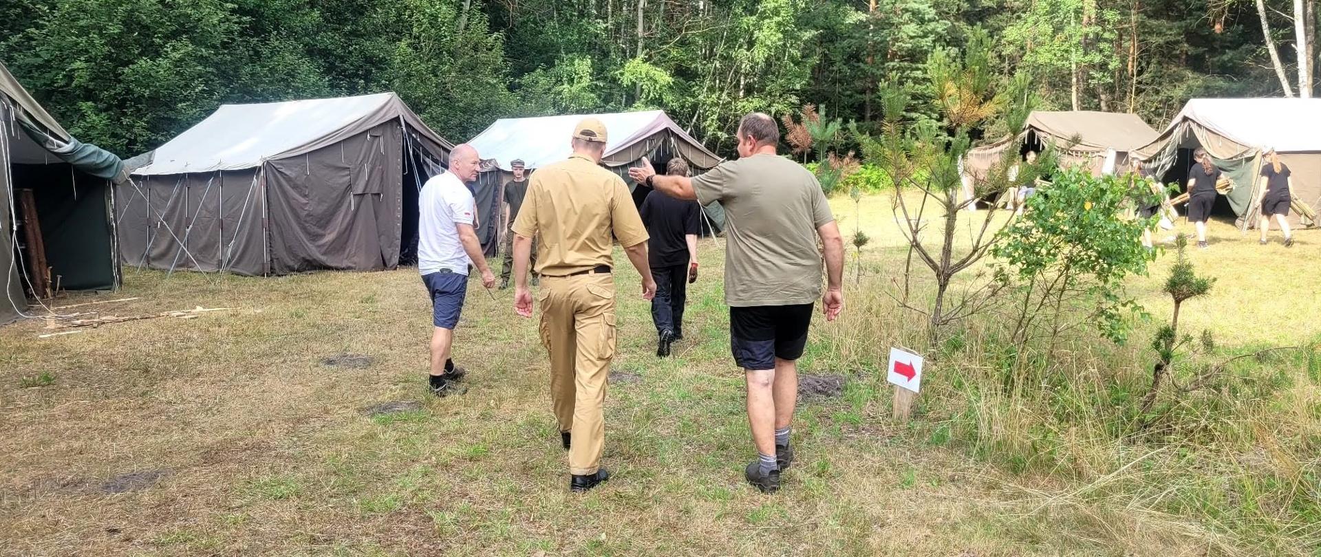 Czterech mężczyzn, w tym jeden w piaskowym mundurze i czapce z daszkiem, idzie tyłem do obiektywu przez trawiastą polanę, na której rozstawione są duże, ciemne namioty wojskowe w sąsiedztwie gęstego, zielonego lasu.