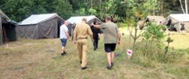 Czterech mężczyzn, w tym jeden w piaskowym mundurze i czapce z daszkiem, idzie tyłem do obiektywu przez trawiastą polanę, na której rozstawione są duże, ciemne namioty wojskowe w sąsiedztwie gęstego, zielonego lasu.
