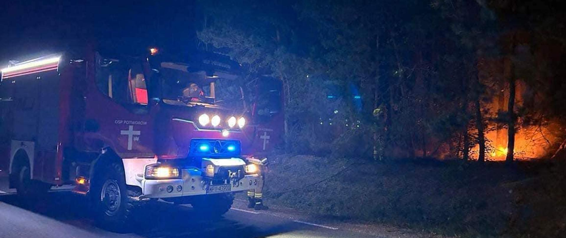 Szerokie, ciemne, nocne ujęcie przedstawia czerwony wóz strażacki zaparkowany na poboczu utwardzonej drogi. Jego niebieskie światła awaryjne migają, oświetlając przód pojazdu i niewielki fragment drogi. W tle, po prawej stronie wozu, intensywnie płonie pożar lasu, z jasnopomarańczowymi płomieniami widocznymi między drzewami. Reszta lasu jest ciemna, zarysowana na tle blasku ognia. Scena przedstawia straż pożarną gaszącą pożar lasu w nocy.