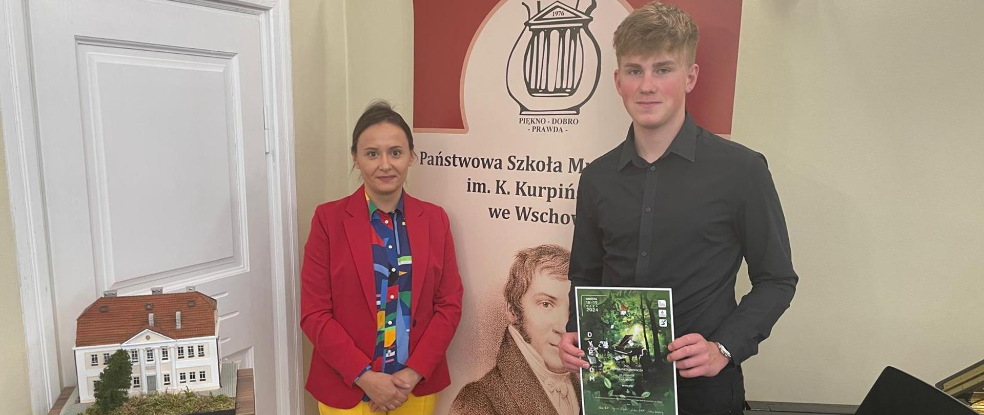 Pani Marta Sadowska i Piotr Fiedorowicz stojący przy fortepianie w auli szkoły. Uczeń trzyma w dłoniach dyplom.