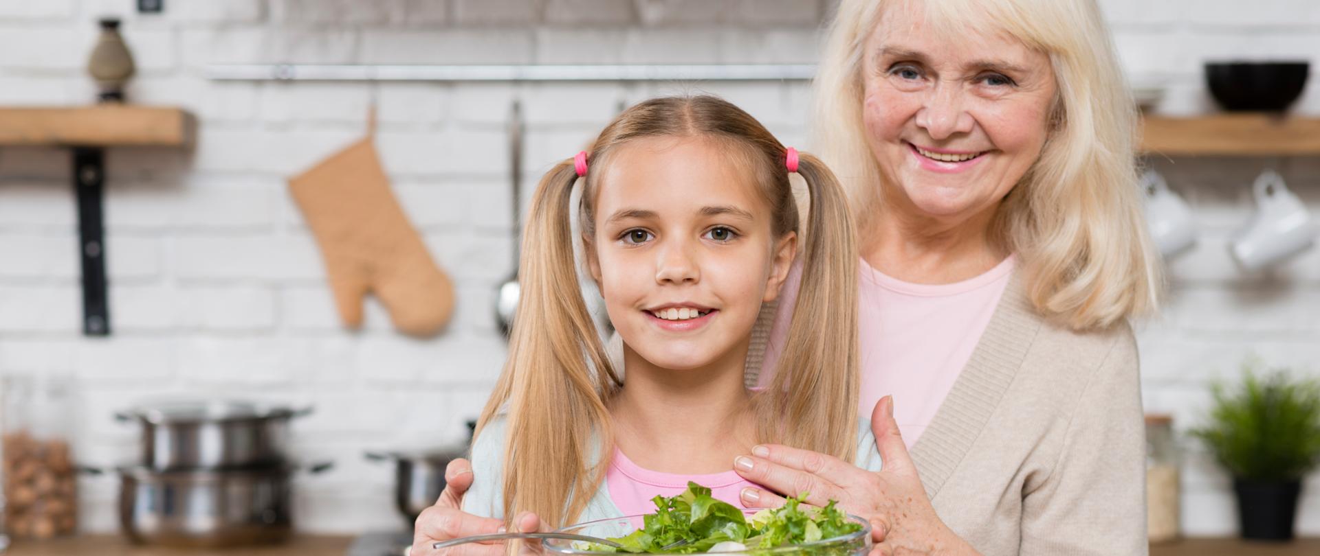 Starsza pani - babcia obejmuje dziewczynkę - wnuczkę, która trzyma w dłoni miskę z sałatką