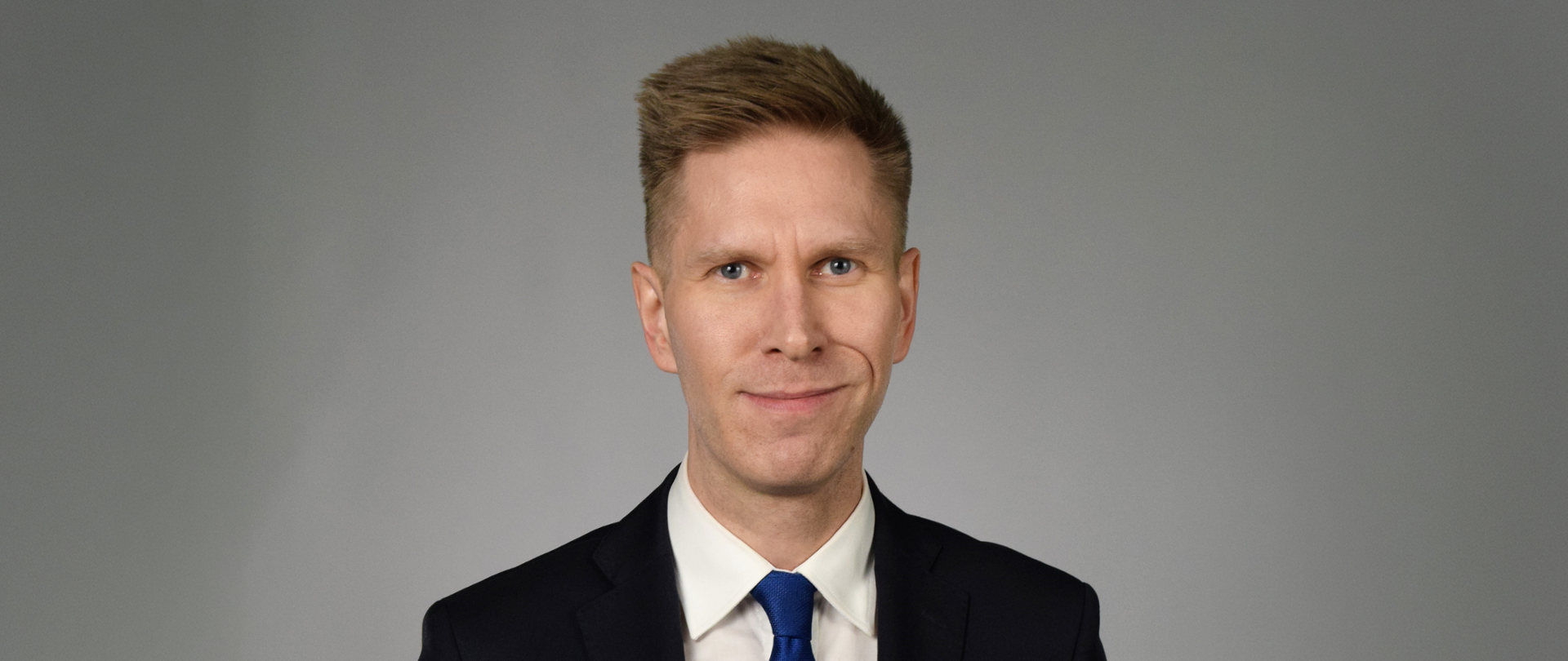 Man in a black suit with a blue tie on a grey background.
