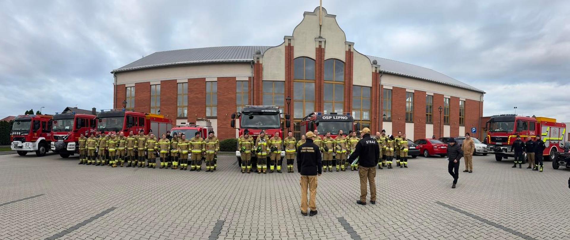 Strażacy stoją w dwuszeregu na tle samochodów strażackich i budynku ratusza.