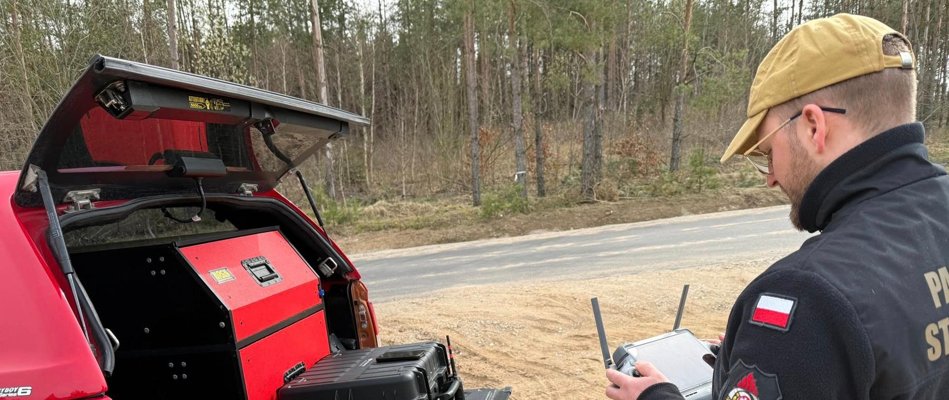 Strażak stojący na poboczu drogi trzyma w dłoniach pilot do sterowania dronem. Mężczyzna stoi przed samochodem specjalnym straży pożarnej. Samochód typu pickup z otwartą klapą bagażnika. Na otwartej klapie znajduje się walizka do transportu drona oraz radiotelefon noszony.