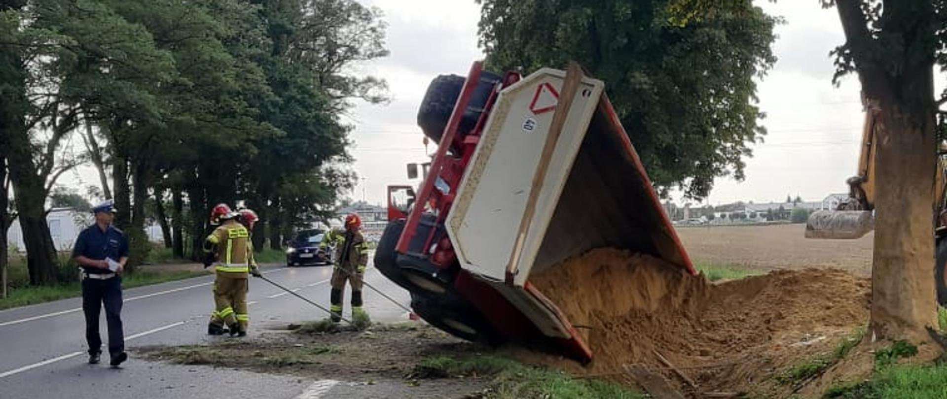 Przewrócona przyczepa z piaskiem w rowie. Obok pracujący strażacy oraz policjanci. 