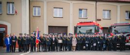 Zdjęcie przedstawia dużą grupę strażaków oraz osób towarzyszących, ustawionych do pamiątkowego zdjęcia przed budynkiem straży pożarnej. W tle widoczne są dwa czerwone wozy strażackie marki Scania, a na budynku można zauważyć polską flagę.
Osoby znajdujące się na zdjęciu są ubrane w galowe mundury strażackie, część z nich ma medale i odznaczenia. Na środku widnieje poczet sztandarowy z biało-czerwonym sztandarem. Po lewej stronie stoją także osoby w cywilnych ubraniach, przedstawiciele władz lokalnych i goście honorowi.
Zdjęcie zostało wykonane z okazji uroczystości Dnia Strażaka w Komendzie Powiatowej PSP w Będzinie. Atmosfera jest oficjalna i podniosła.
