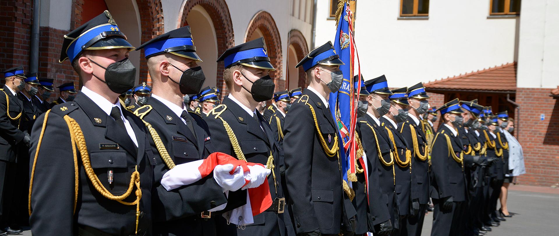 Strażacy stojący w ugrupowaniu rozwiniętym na placu JRG1 w Bydgoszczy, wszyscy mundury wyjściowe i maseczki na twarzy