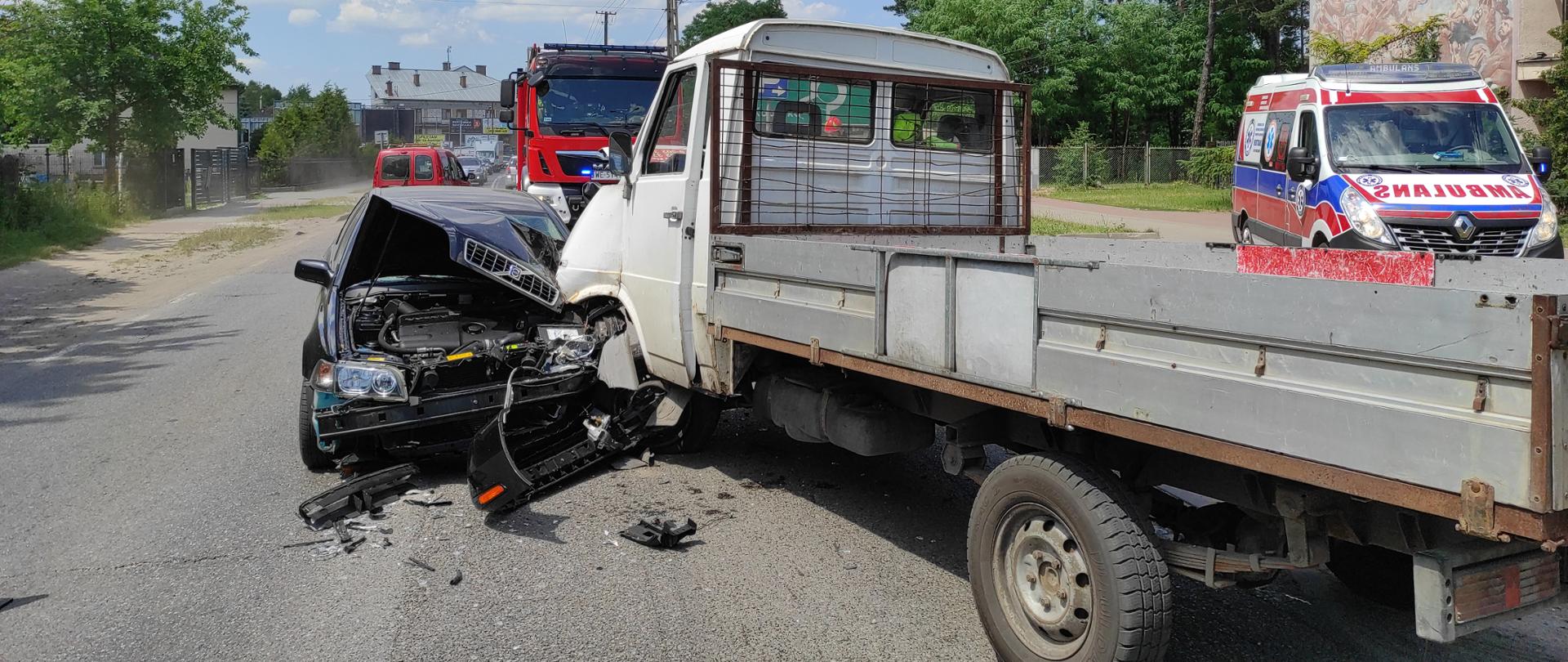 Na środku zdjęcia widać drogę asfaltową, na której znajdują się dwa rozbite samochody: jeden osobowy koloru czarnego, drugi dostawczy koloru białego. Pojazdy zderzyły się czołowo, widoczne są zniszczone przednie części nadwozia. Za miejscem wypadku znajduje się czerwony samochód straży pożarnej, zaś w oddali widoczny jest rząd samochodów oczekujących na przejazd. Po prawej stronie zdjęcia znajduje się karetka pogotowia ratunkowego. Przy rozbitych pojazdach, na asfalcie rozrzucone są połamane elementy samochodów.