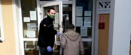 Strażak Ochotniczej Straży Pożarnej w maseczce ochronnej i rękawicach lateksowych otwiera drzwi do przychodni, wprowadzając tam starszą panią, która udaje się do punktu szczepień