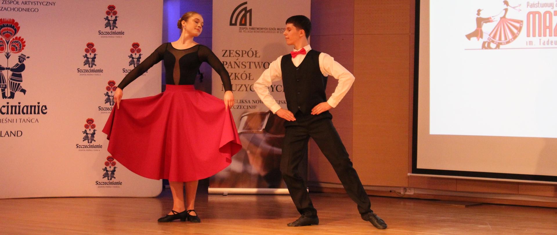 zdjęcie przedstawia parę wykonawców tańczących na scenie sali koncertowej, w tle baner wydarzenia szkoły oraz ekran multimedialny