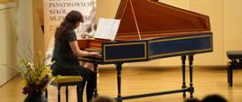 zdjęcie przedstawia uczennicę grającą na klawesynie na scenie sali koncertowej, w tle baner z logo szkoły