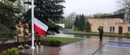 Przed budynkiem Komendy uroczystość Dnia Flagi. Trzech strażaków w umundurowaniu specjalnym koloru piaskowego i hełmach czerwonych, mocują flagę Polski do masztu flagowego. W oddali stoi strażak na baczność oddając honor. Dzień pochmurny, deszczowy.