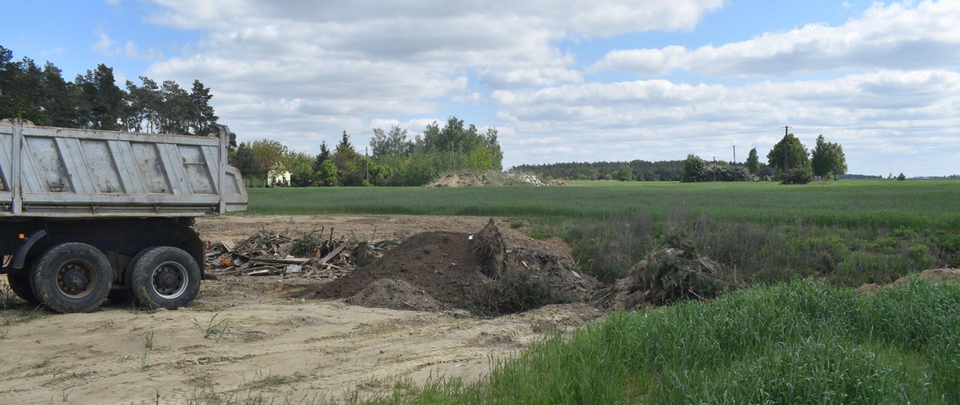 Na porośniętym trawą płaskim terenie znajduje się brązowe pryzmy zawierające zmieszane odpady. Po lewej stronie widać tył samochodu ciężarowego, z którego te odpady zostały zsypane.