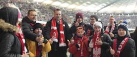 Na murawie stadionu stoi minister Czarnek w czarnej kurtce i biało-czerwonym szaliku, obok niego grupa dzieci w kolorowych kurtkach i zimowych czapkach.