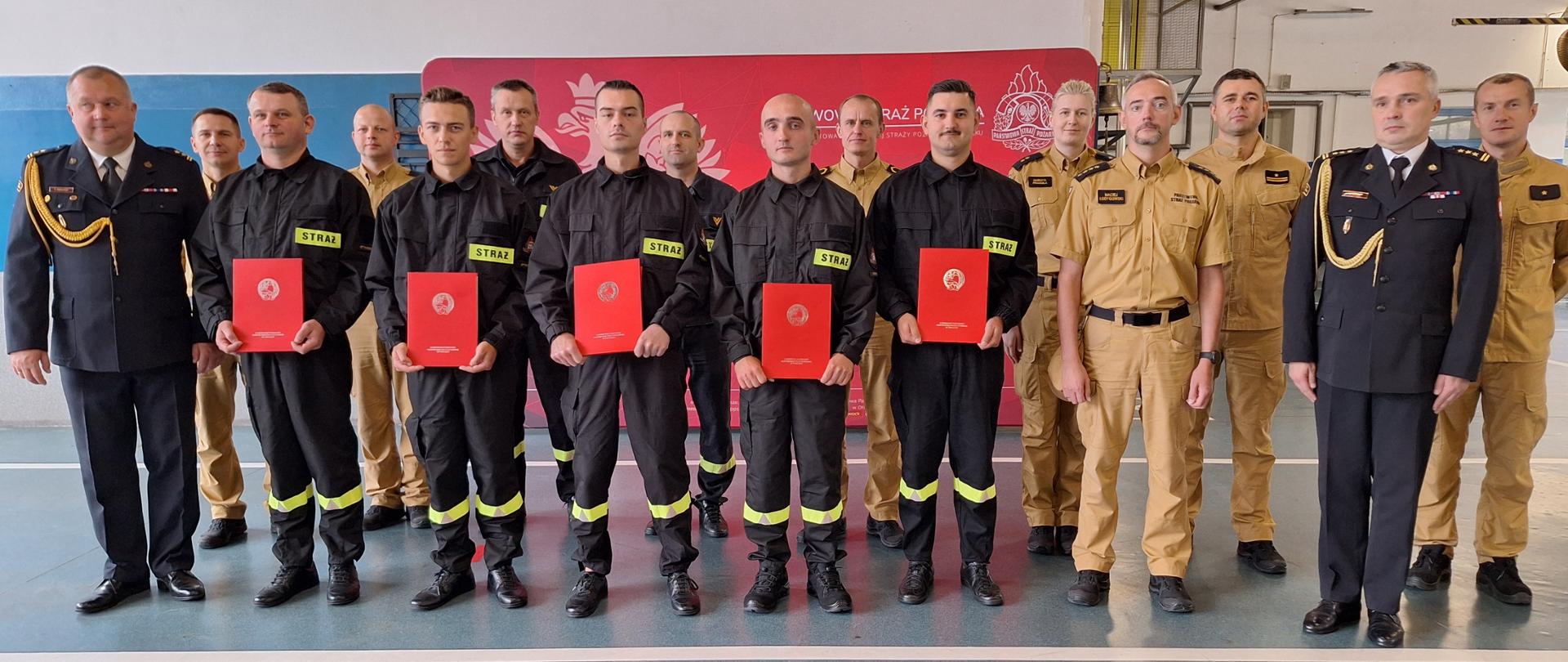 Na zdjęciu widzimy grupowe zdjęcie wykonane po zakończeniu uroczystości ślubowania.
Na pierwszym planie stoi siedmiu mężczyzn ubranych w czarne mundury z napisem „STRAŻ” – każdy z nich trzyma w rękach czerwony dokument z godłem Polski (orzeł biały w koronie). Po bokach grupy znajdują się dwaj oficerowie w galowych mundurach straży pożarnej, z odznaczeniami i złotymi sznurami naramiennymi, co sugeruje wysokie stanowiska służbowe.
W tle stoją pozostali strażacy, głównie w beżowych (piaskowych) mundurach roboczych, typowych dla Państwowej Straży Pożarnej. Za grupą znajduje się czerwony baner z logotypem Komendy Powiatowej Państwowej Straży Pożarnej oraz grafiką strażackiego.
Jest to zakończenie ślubowania nowych funkcjonariuszy. Atmosfera jest uroczysta i oficjalna, a uśmiechy oraz postawa uczestników sugerują dumę i satysfakcję z osiągnięcia.
