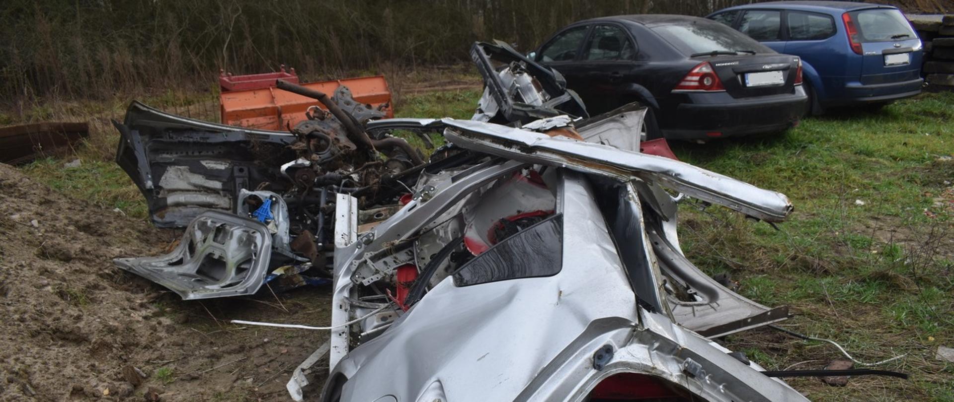 Porozrzucane, zniszczone części samochodowe i elementy karoserii leżą na ziemi na nieuporządkowanym terenie. Obok, na nieutwardzonym podłożu, stoją dwa zaparkowane auta osobowe.