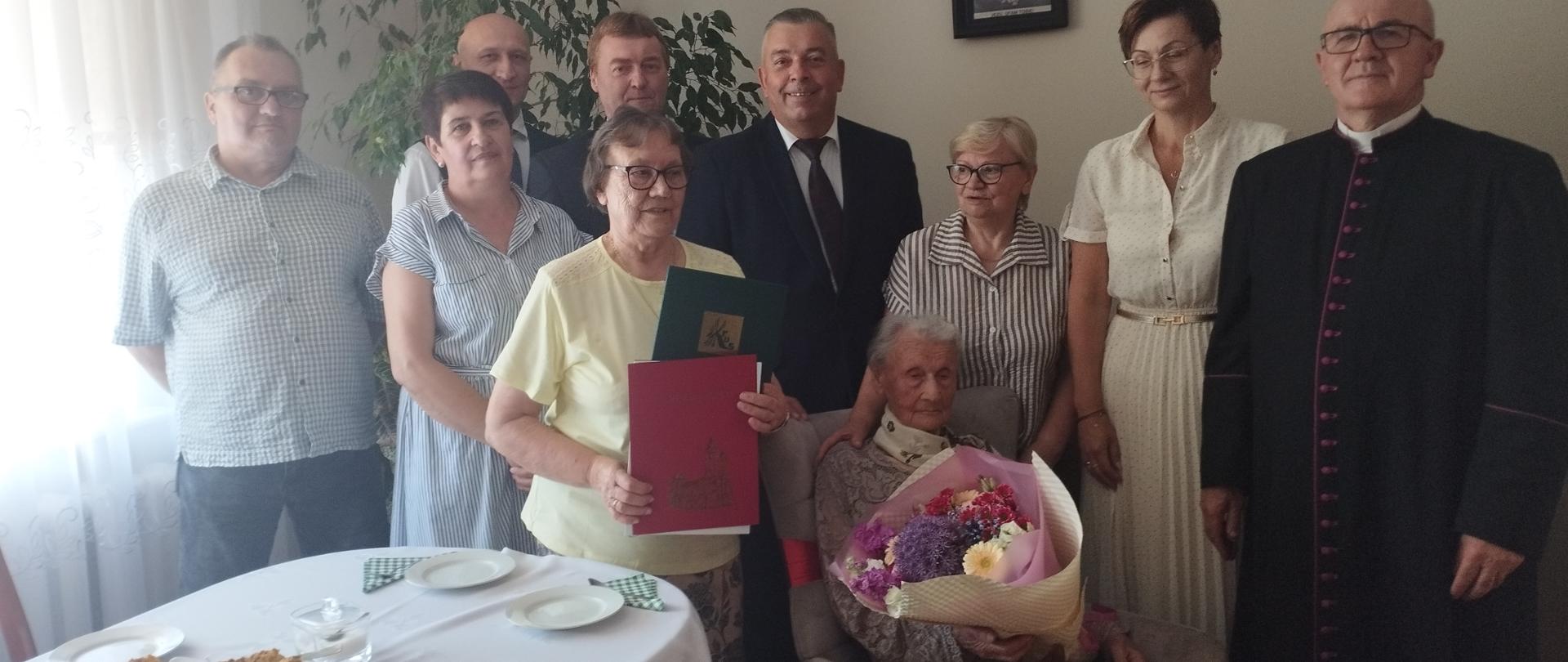 W jasno pomalowanym wnętrzu pokoju przy stole odświętnie nakrytym białym obrusem, na szarym fotelu siedzi starsza stuletnia kobieta trzymająca w rękach bukiet kolorowych kwiatów. W jej otoczeniu obok i lekko z tyłu stoją elegancko ubrane kobiety – córki oraz mężczyźni w garniturach. W gronie obecnych osób postać mężczyzny – proboszcza parafii w czarnej, długiej sutannie. Wszyscy są przejęci chwilą i uśmiechają się