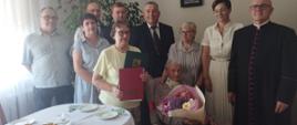 W jasno pomalowanym wnętrzu pokoju przy stole odświętnie nakrytym białym obrusem, na szarym fotelu siedzi starsza stuletnia kobieta trzymająca w rękach bukiet kolorowych kwiatów. W jej otoczeniu obok i lekko z tyłu stoją elegancko ubrane kobiety – córki oraz mężczyźni w garniturach. W gronie obecnych osób postać mężczyzny – proboszcza parafii w czarnej, długiej sutannie. Wszyscy są przejęci chwilą i uśmiechają się
