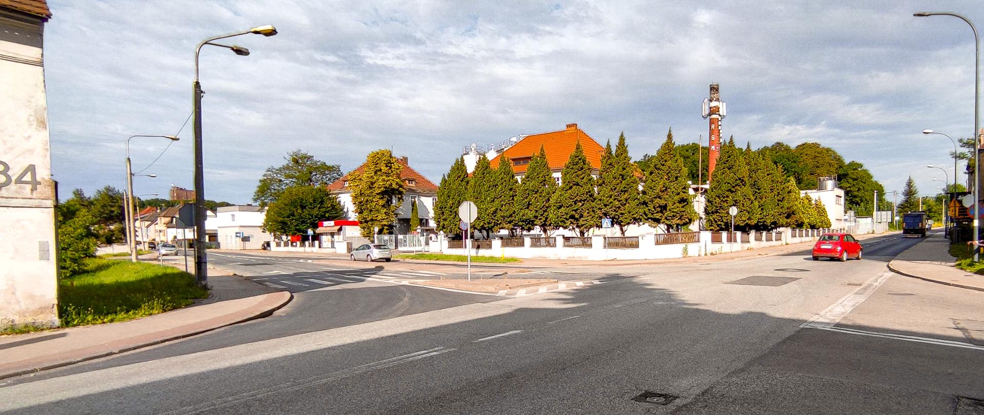 Na zdjęciu widoczna jest ulica na skrzyżowaniu, po której porusza się czerwony samochód, jadący prawą stroną jezdni. W tle znajdują się budynki, w tym jeden z czerwonym dachem, oraz drzewa. Wzdłuż dróg rozmieszczone są latarnie uliczne i znaki drogowe. Niebo jest częściowo zachmurzone.