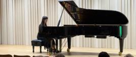 Dziewczyna brunetka gra recital fortepianowy na dużym czarnym fortepianie koncertowym na scenie sali koncertowej. Pianistka ubrana jest na czarno i siedzi przy instrumencie ustawionym na jasnej drewnianej podłodze, na tle jasnych pionowych paneli akustycznych. Na pierwszym planie widoczna jest publiczność siedząca w rzędach sali koncertowej i słuchająca występu w ramach Gdańskiego Festiwalu Muzycznego.