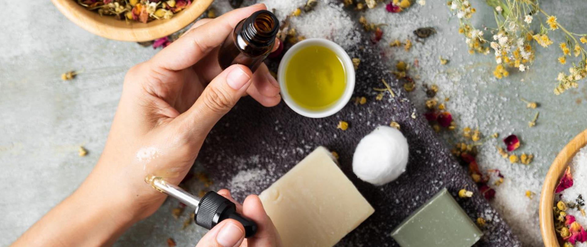 Na zdjęciu znajduje się dłoń trzymająca buteleczkę. Druga ręka aplikatorem nakłada kosmetyk. W tle widać mydełka i oliwkę w miseczce.