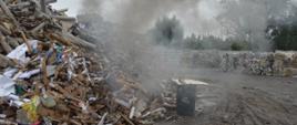 Akcja na składowisku odpadów. Plac, na którym znajdują się pryzmy z odpadami. Nad pryzmami unosi się dym. W tle betonowa ściana i drzewa.