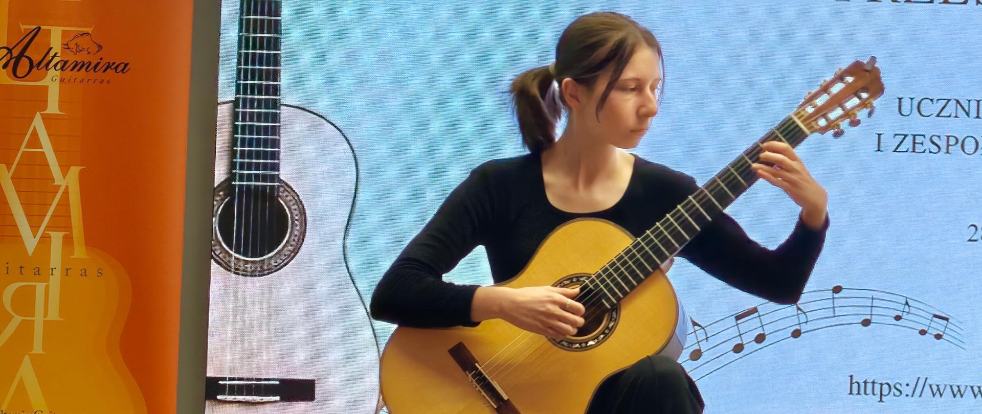 Dziewczynka ubrana na czarno-białe buty lewa noga spoczywa na podnóżku w rękach trzyma gitarę klasyczną na której gra obok stoją dwa krzesła i dwa podnóżki gitarowe.
Z tyłu za nią widać baner reklamowy gitar Altamira. W tle wyświetla się napis Państwowa Szkoła Muzyczna I stopnia im. Zygmunta Mycielskiego w Strzyżowie. Poniżej XVI Makroregionalne Przesłuchania uczniów klas gitary i zespołów gitarowych 28 maja 2026 roku.