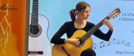 Dziewczynka ubrana na czarno w rękach trzyma gitarę klasyczną na której gra.
Z tyłu za nią widać baner reklamowy gitar i biały ekran.
