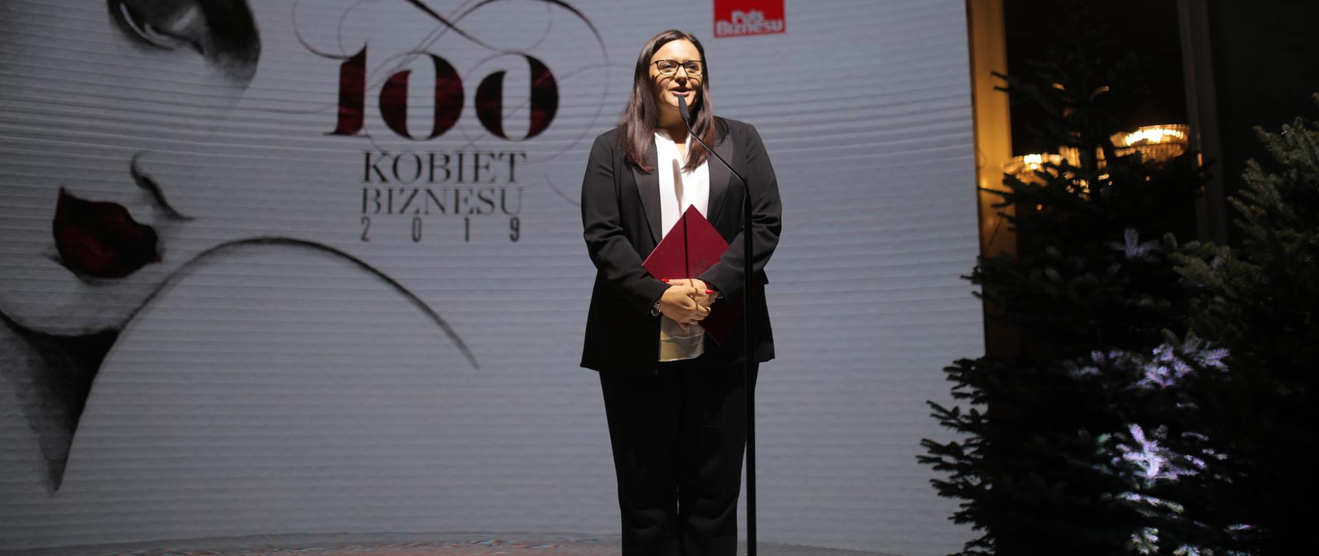 Na tle białej ścianki z napisem „100 Kobiet Biznesu” stoi prze mikrofonem Małgorzata Jarosińska-Jedynak. Ubrana jest w czarne spodnie i marynarkę oraz białą bluzkę. Po prawej stronie widać choinki.